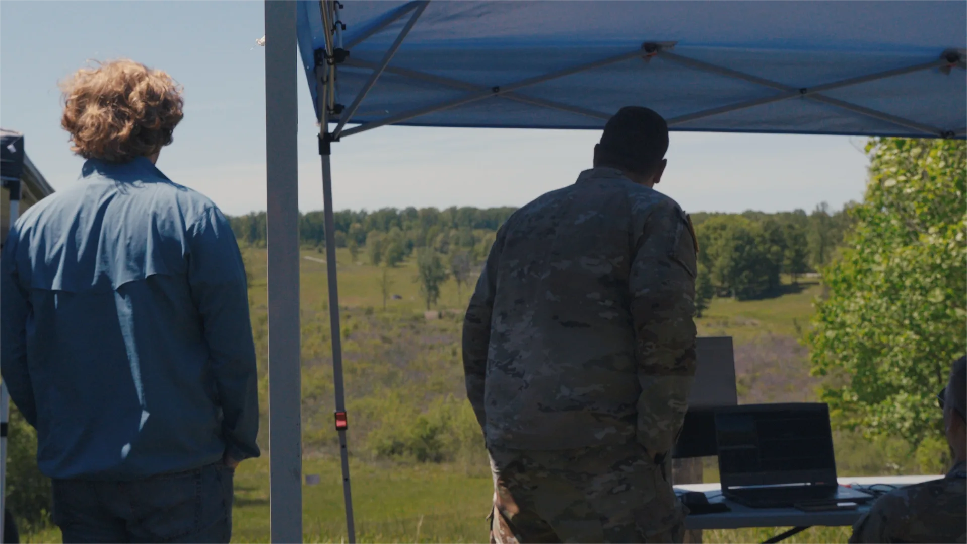 A civilian engineer and a service member observe a daytime field demonstration from a test station